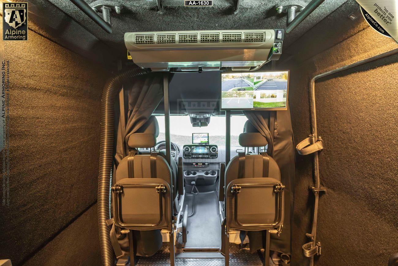 Interior view of an armored Pointer Van showing two front seats, monitors above the windshield displaying external views, and various equipment in the surrounding area.