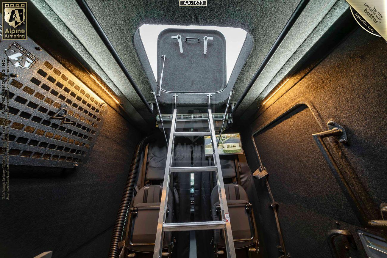 Interior view of a compact, armored Mercedes-Benz Pointer Van showing a secured door hatch on the ceiling with a metal ladder leading up to it. There are two seats and various compartments along the sides.