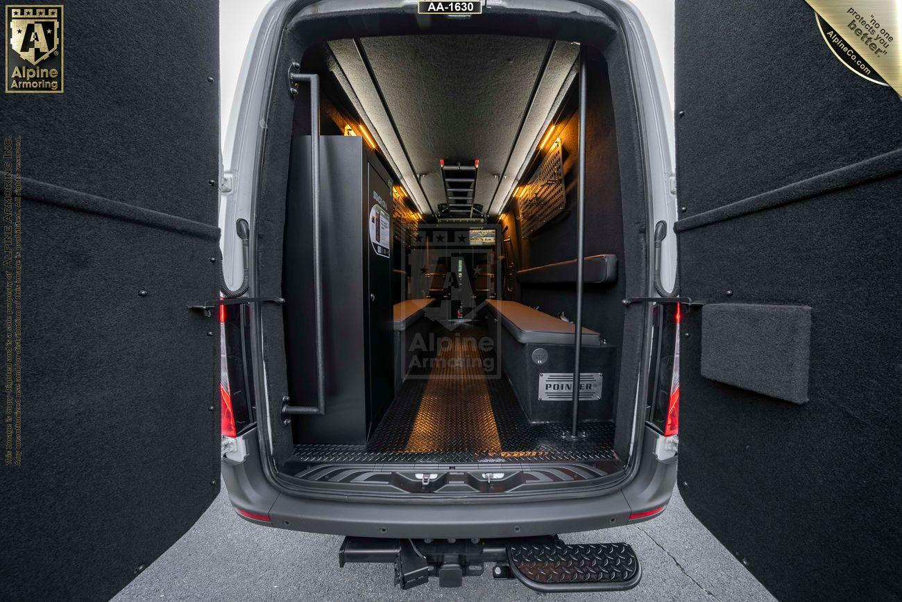 Interior view of an armored Pointer pointer based on Mercedes Sprinter 3500XD van with metal flooring, padded walls, and secured storage compartments. The open rear doors reveal the spacious, fortified space.