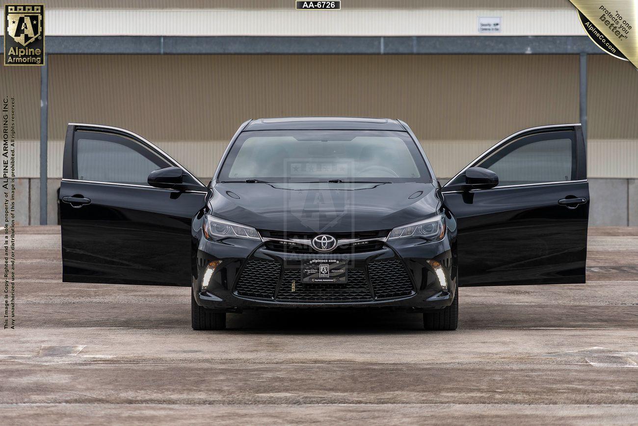 A black Toyota Camry with both front doors open, parked in a lot in front of a warehouse.