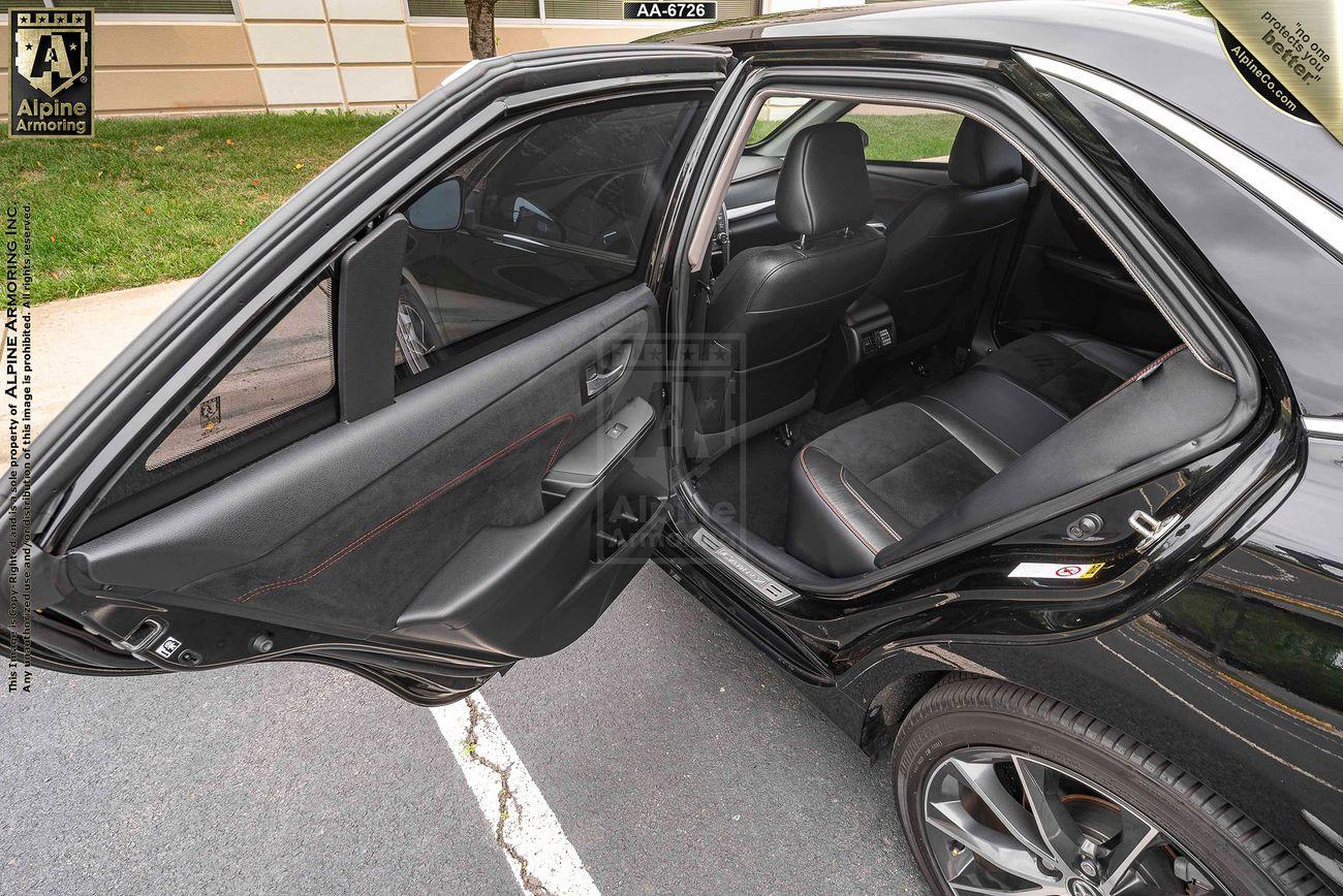 A black armored Toyota Camry sedan with its rear door open, showing the black leather interior and back seats. The grass and part of a building are visible in the background.