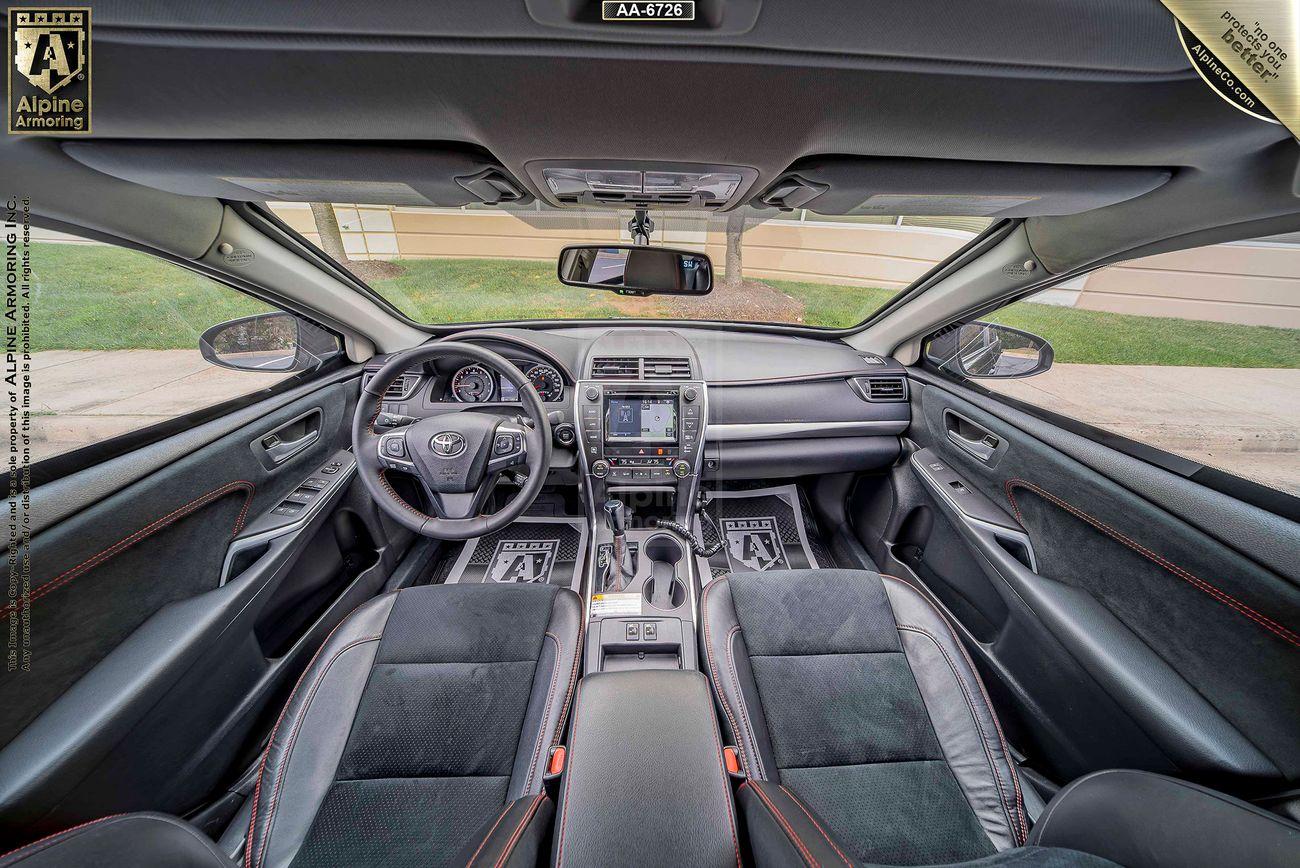 Wide-angle view of a Toyota Camry's interior, showing two front seats, a dashboard with multiple controls, and a steering wheel adorned with the Toyota logo. Sunroof overhead and a grassy exterior visible.