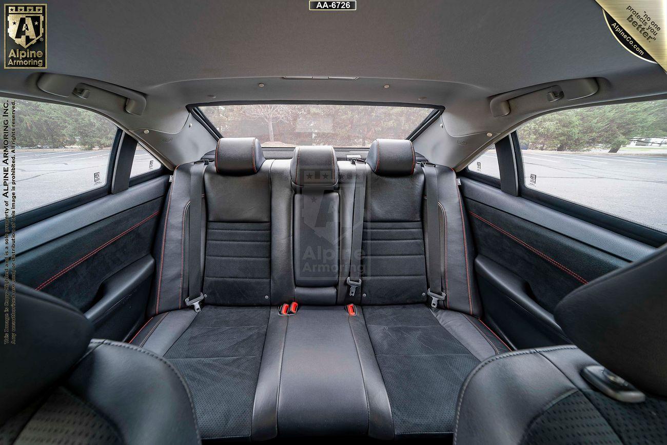 Interior view of a bulletproof Toyota Camry showcasing the rear passenger seats with black leather upholstery and red stitching. The back window and partial rearview of the exterior are visible through the windows.