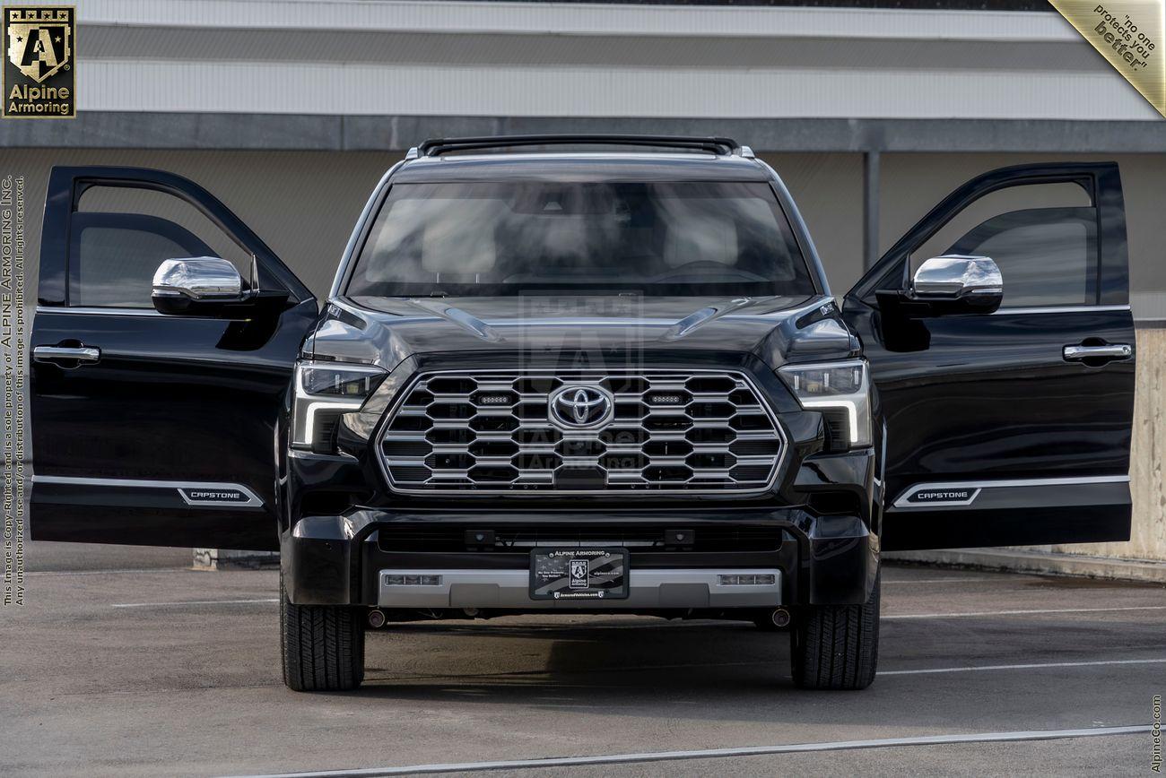 The front view of a white armored Toyota Sequoia Capstone from Alpine Armoring parked in front of a modern building with large glass windows.