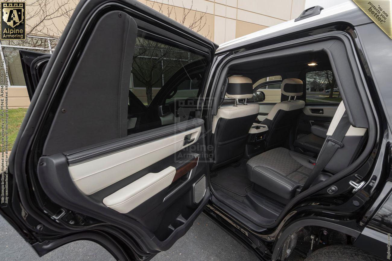 The interior of a  A white armored Toyota Sequoia Capstone from Alpine Almoring parked near a building with large windows.