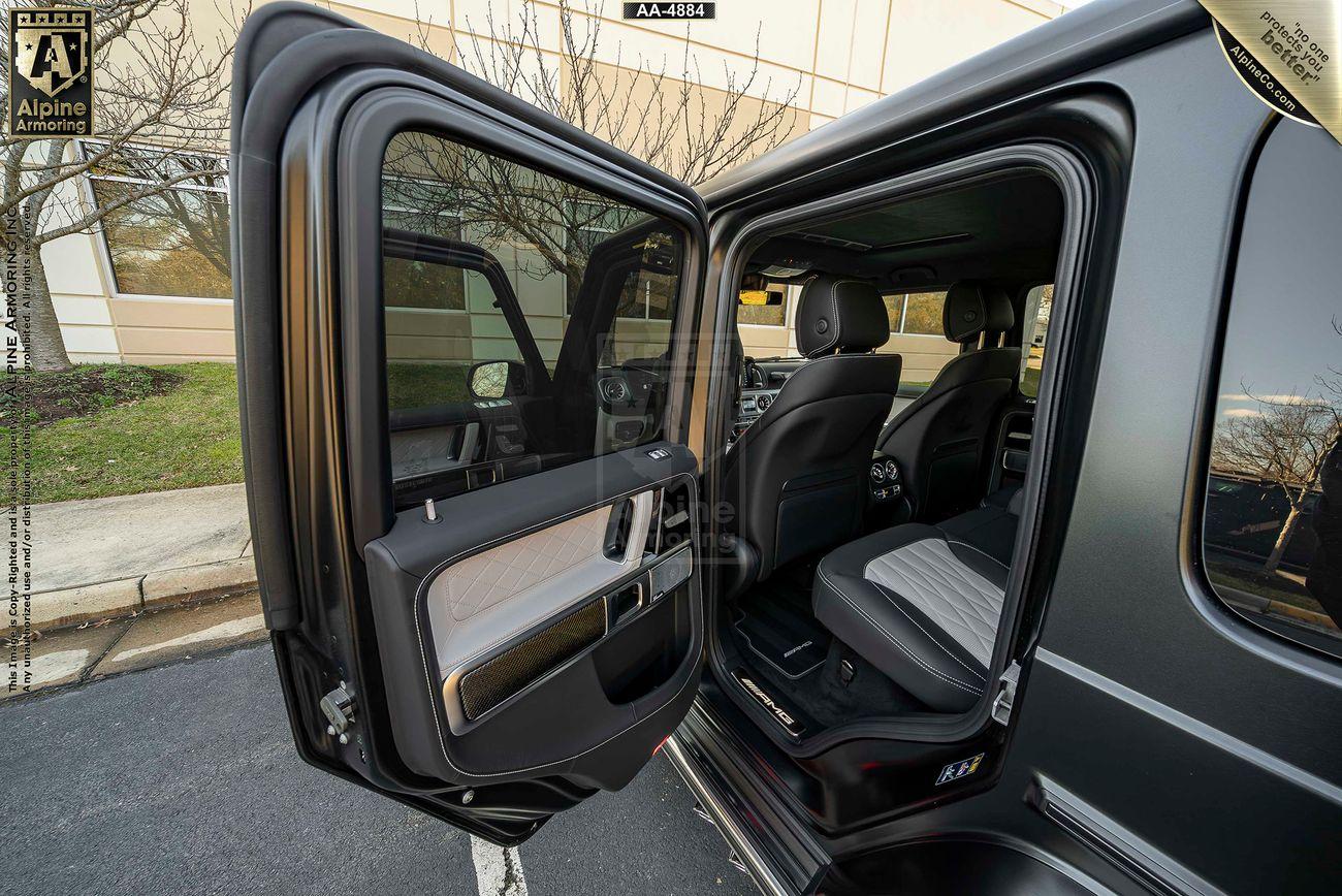 An open rear passenger door of a black armored Mercedes-Benz G63 AMG SUV showing the bulletproof glass in the door, a view of the interior seats and a building in the background.