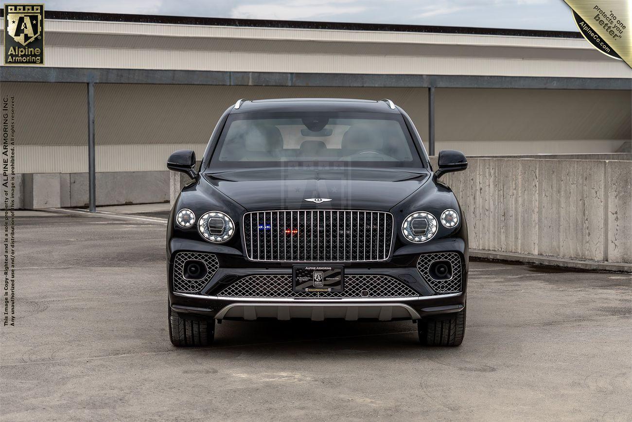 A front view of a black Bentley Bentayga SUV parked in an outdoor area, with the Alpine Armoring logo visible on the upper left corner. The car displays distinctive headlights and a prominent grille.