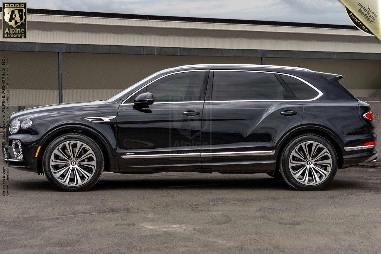 A black Bentley Bentayga luxury SUV is parked on a concrete surface in front of a building, viewed from the side. The vehicle has tinted windows, intricate alloy wheels, and a sleek, modern design.