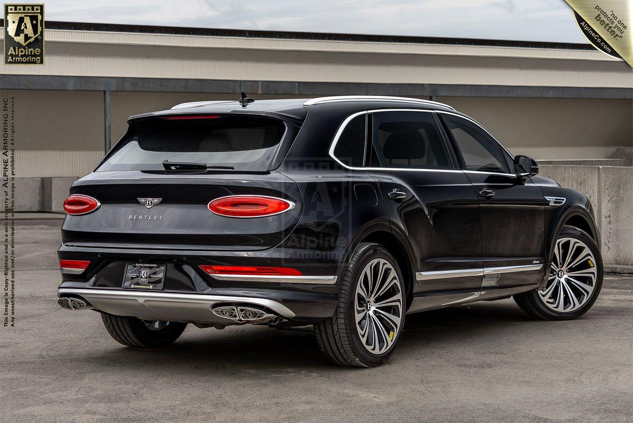 A black armored Bentley Bentayga SUV is parked outdoors in an empty concrete area. The vehicle has tinted windows, chrome detailing, and dual exhaust pipes. A banner on the top right reads "Alpine Armoring.