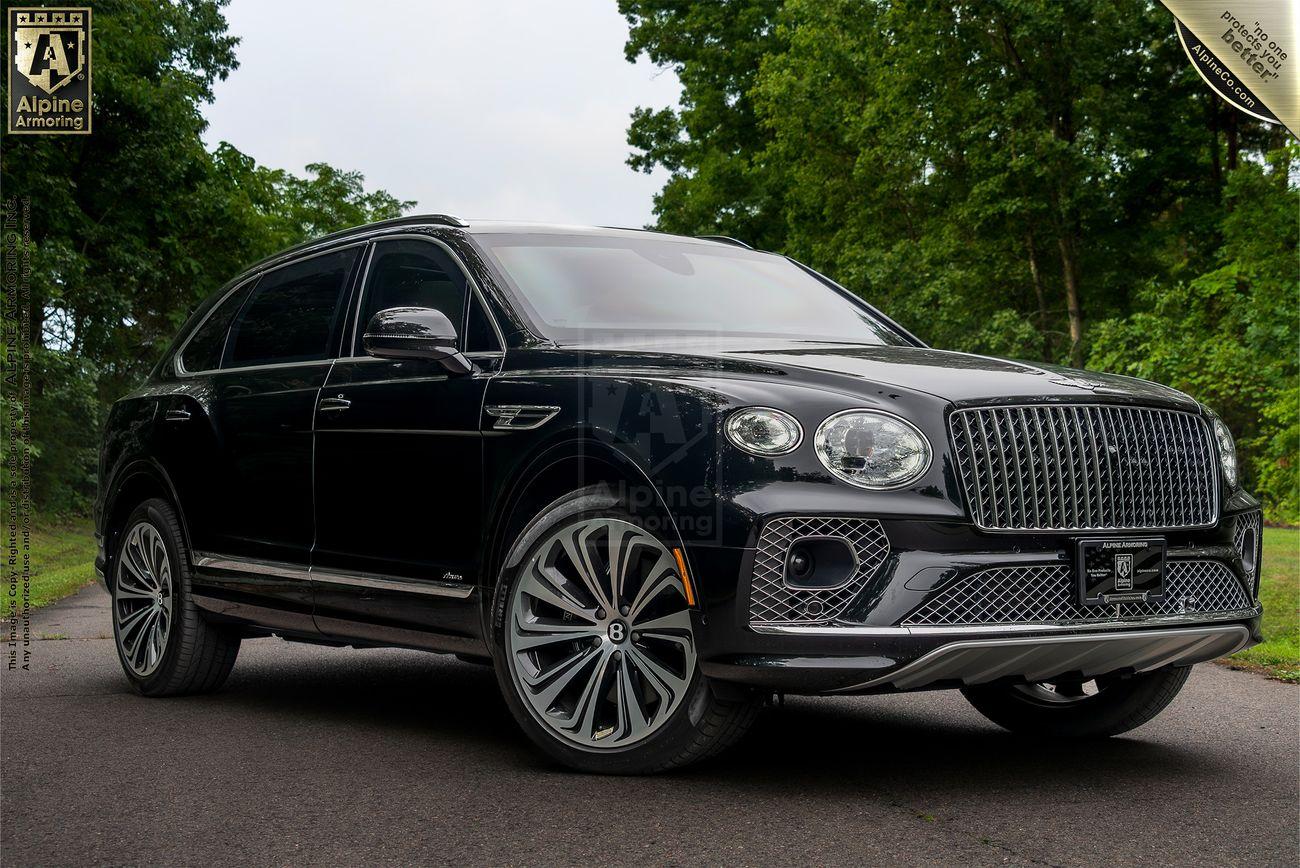 A black luxury Bentley Bentayga SUV is parked on a paved road with trees in the background. The vehicle features large alloy wheels and a distinctive grille.