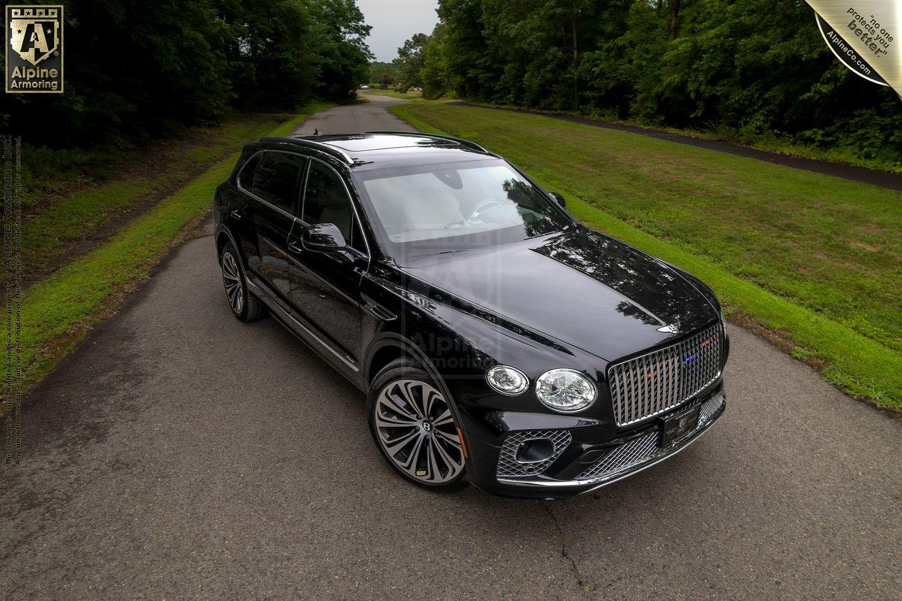 A black SUV is parked on a paved road surrounded by greenery. The badge on its front grille indicates it's a Bentley Bentayga.