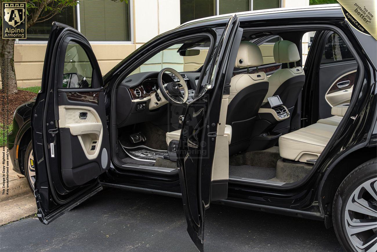 A black armored Bentley Bentayga SUV with both front and rear passenger-side doors open, revealing a beige leather interior. The vehicle is parked on a paved area in front of a building with beige and brown walls.