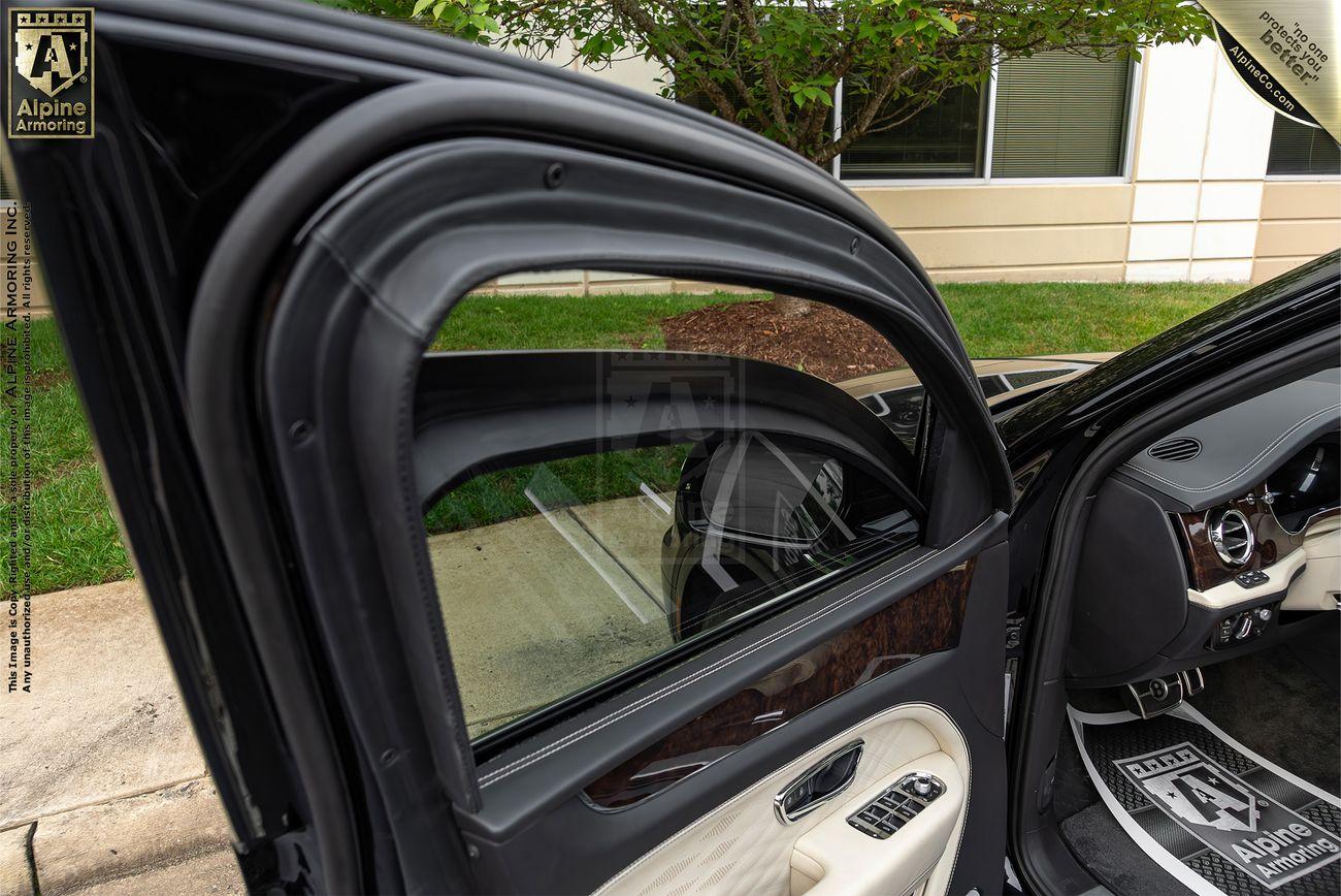 The open rear door of an armored Bentley Bentayga with tinted, bulletproof windows and an Alpine logo. The door interior features light-colored upholstery, window controls, and a "Bentley" watermark.