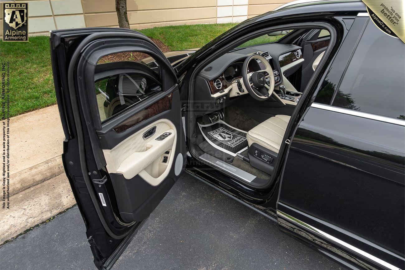 An open car door on an armored Bentley Bentayga revealing the interior of a luxury vehicle with white leather seats, a detailed dashboard, and a "Bentley Motors" logo on the steering wheel.