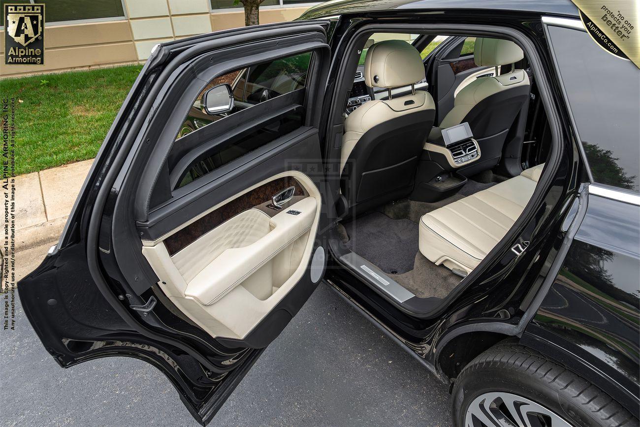 A backseat view of a black armored Bentley Bentayga with beige leather interior and an open rear passenger door. The car is parked near a building.