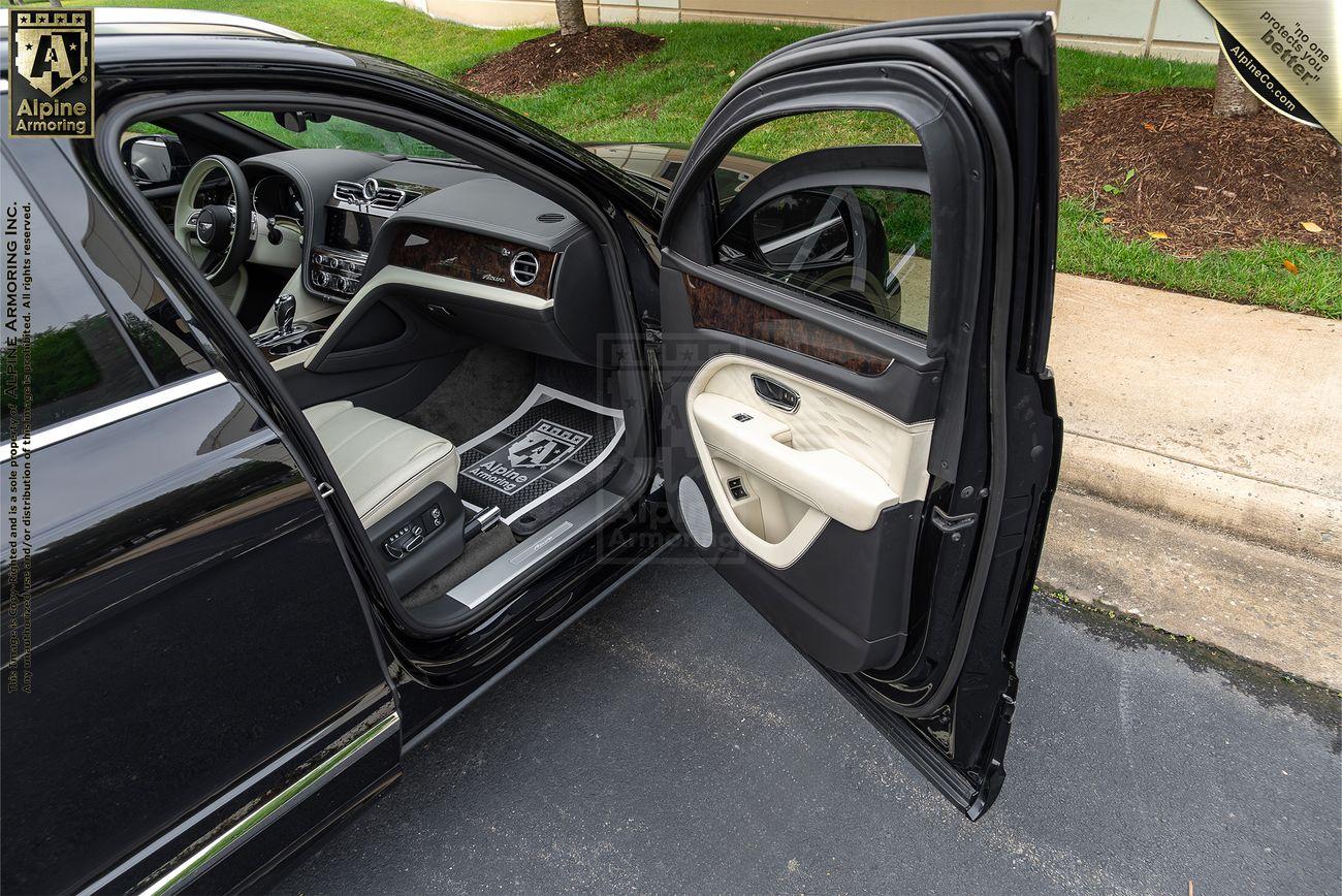 Bentley Bentayga luxury black sedan with its rear passenger door open, revealing a premium interior featuring cream leather seats and a wooden dashboard trim.