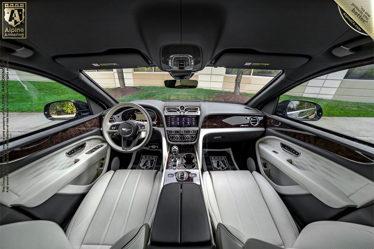 Interior view of an armored Bentley Bentayga luxury SUV with a modern dashboard, leather seats, and wooden accents. The front windshield offers a view of a lawn and wall outside. Branding badges are visible in the corners.