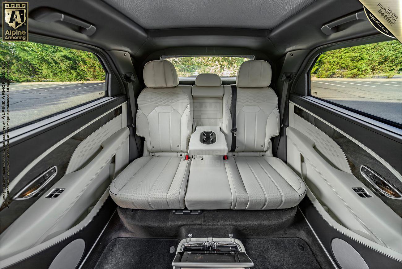 Interior view of a Bentley Bentayga's backseat with three white leather seats, a middle seat cup holder, and black and silver accents. The badge on the top left reads "Alpine Armoring." The image shows the rear cabin.