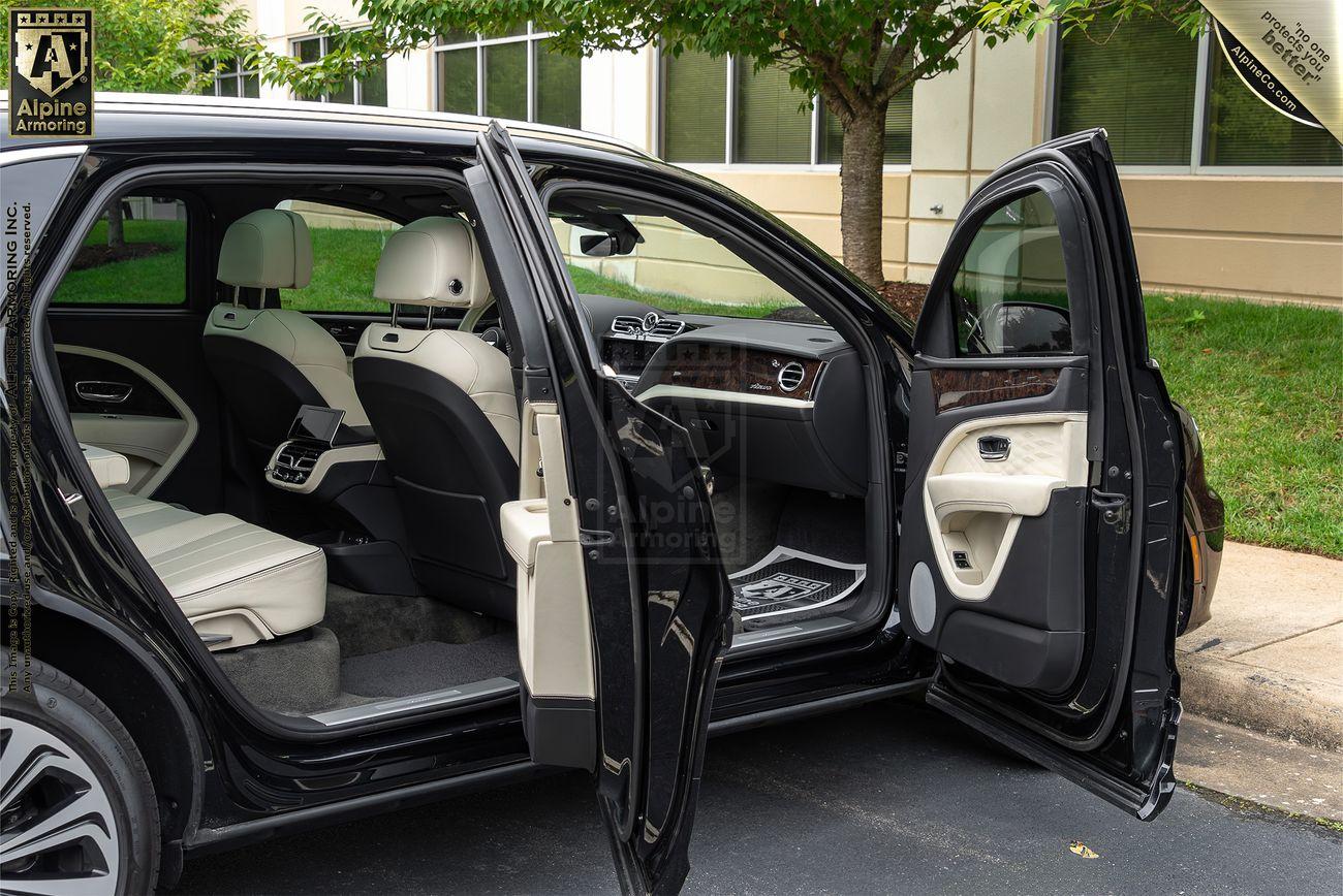 A black armored Bentley Bentayga SUV with its rear passenger-side doors open, showcasing a white leather interior with visible headrests and a center console. The vehicle is parked by a building with windows and trees.