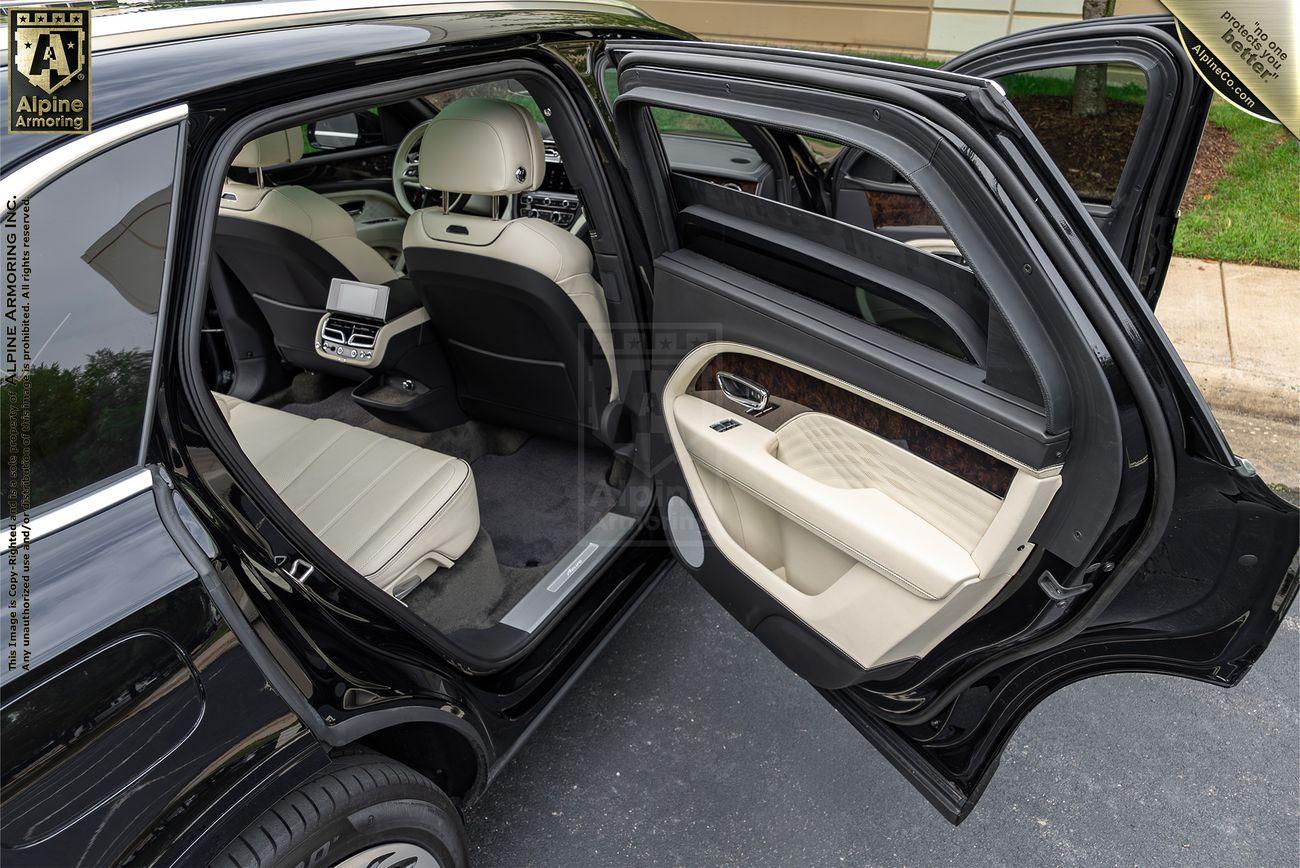 Back door of a black Bentley Bentayga open, revealing beige interior, rear seats, and a tablet displaying a rear camera view.