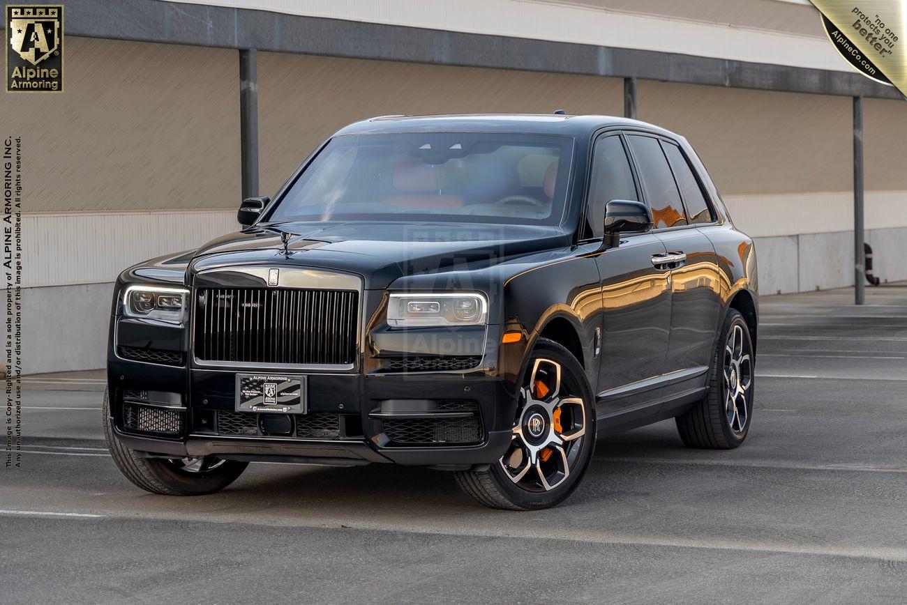 A black armored Rolls-Royce Cullinan SUV parked in an outdoor lot, featuring distinctive orange-accented wheels and a polished exterior under an overcast sky.