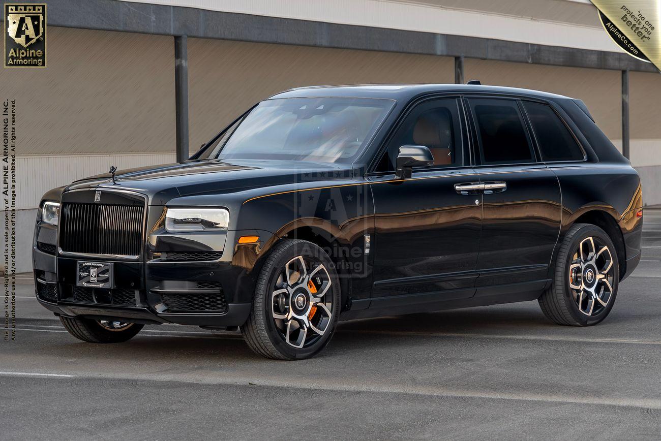 A black bulletproof Rolls-Royce Cullinan SUV is parked in a lot, with custom wheels and detailing from Alpine Armoring Inc. The background displays a beige building.