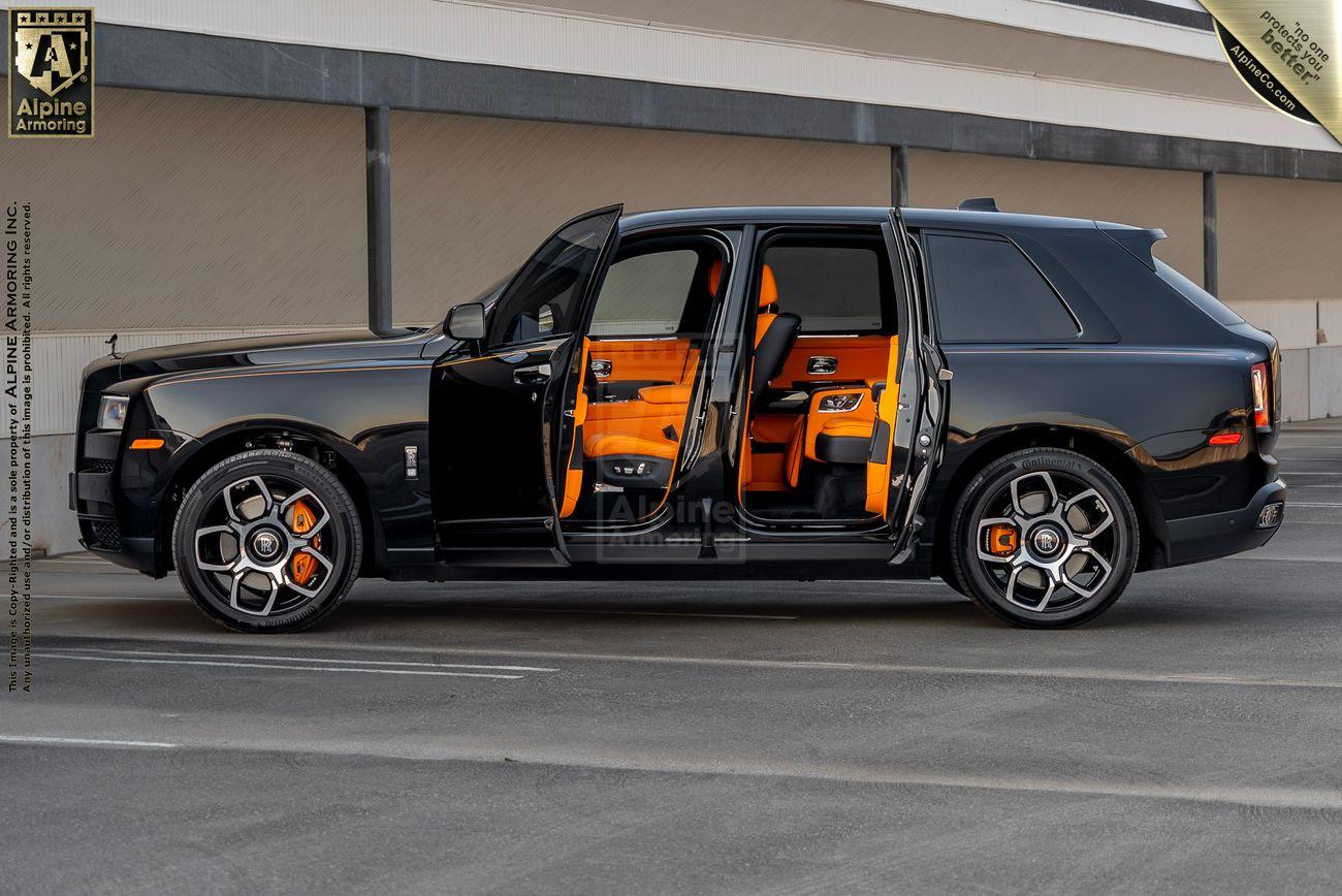 A black armored Rolls-Royce Cullinan SUV with orange interior is parked with all doors open in a deserted area. Background shows a building facade.