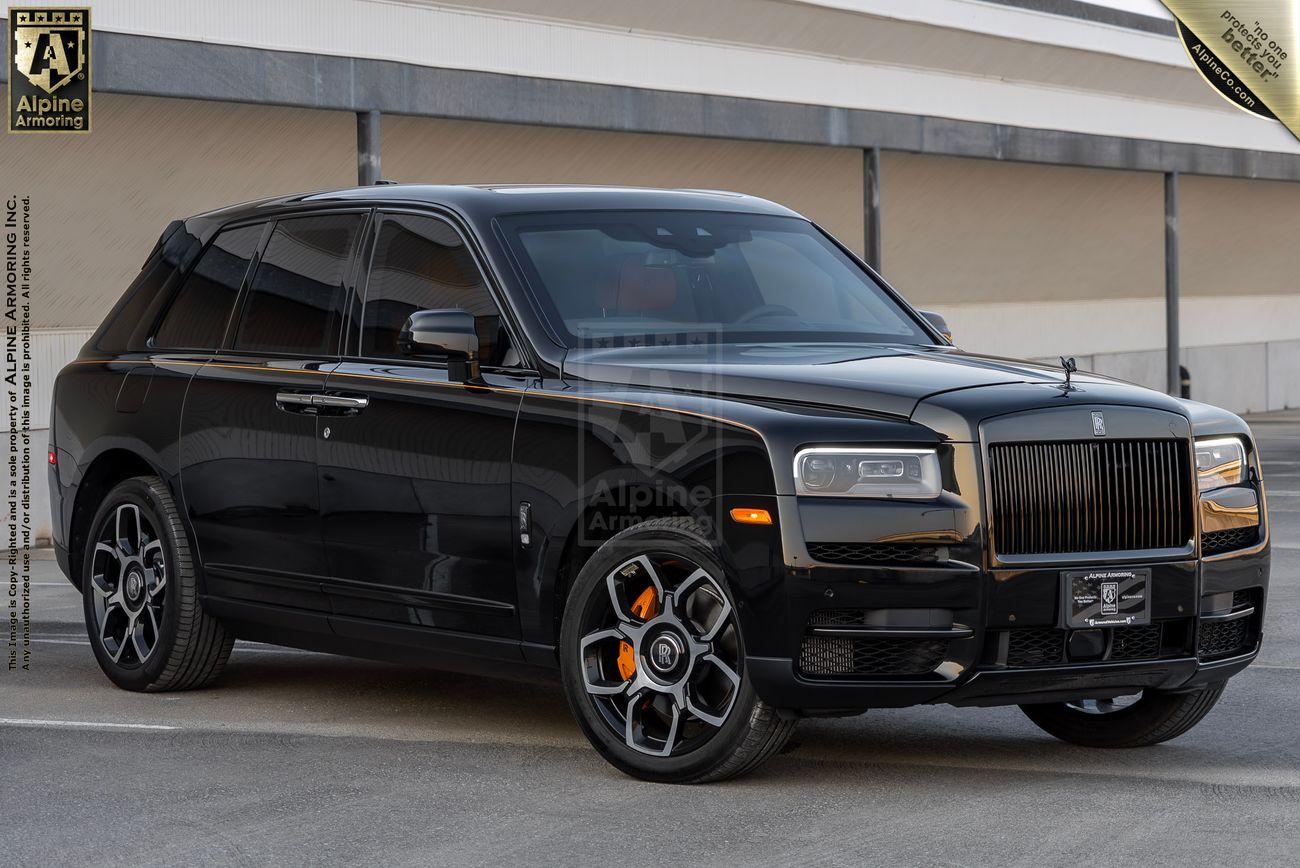 A black armored Rolls-Royce Cullinan SUV is parked on a concrete surface. The vehicle has tinted windows, orange brake calipers, and a polished exterior, with a logo and text on the backdrop and banner overhead.