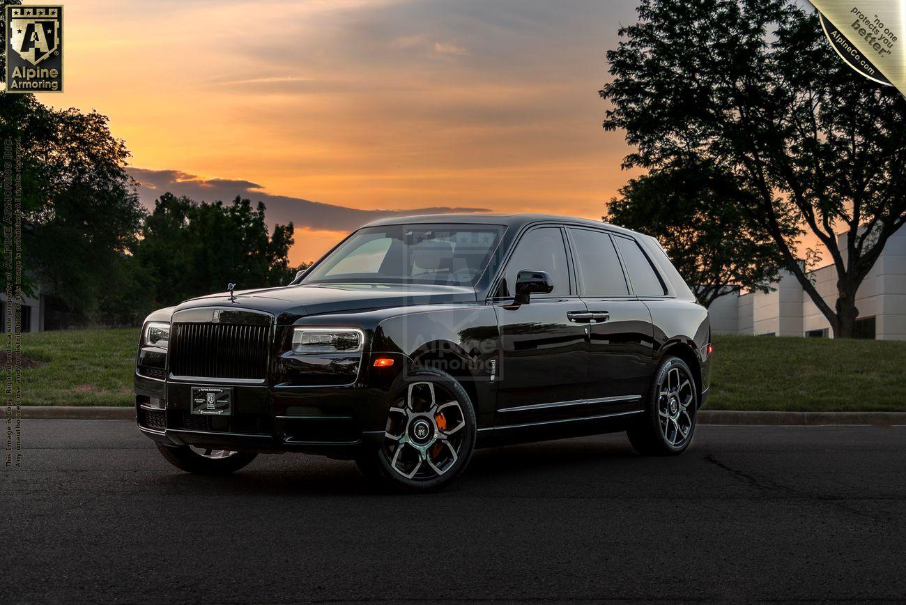 A black armored Rolls-Royce Cullinan SUV is parked on a paved road with trees and a sunset in the background.