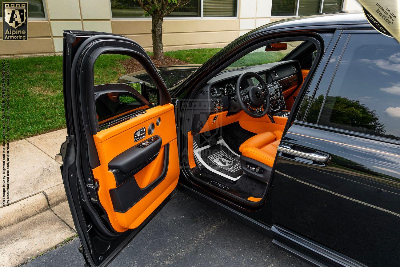 The interior of a armored Rolls-Royce Cullinan SUV with bright orange leather seats. The driver-side door is open, revealing the dashboard and steering wheel. A mat on the floor displays a geometric pattern.