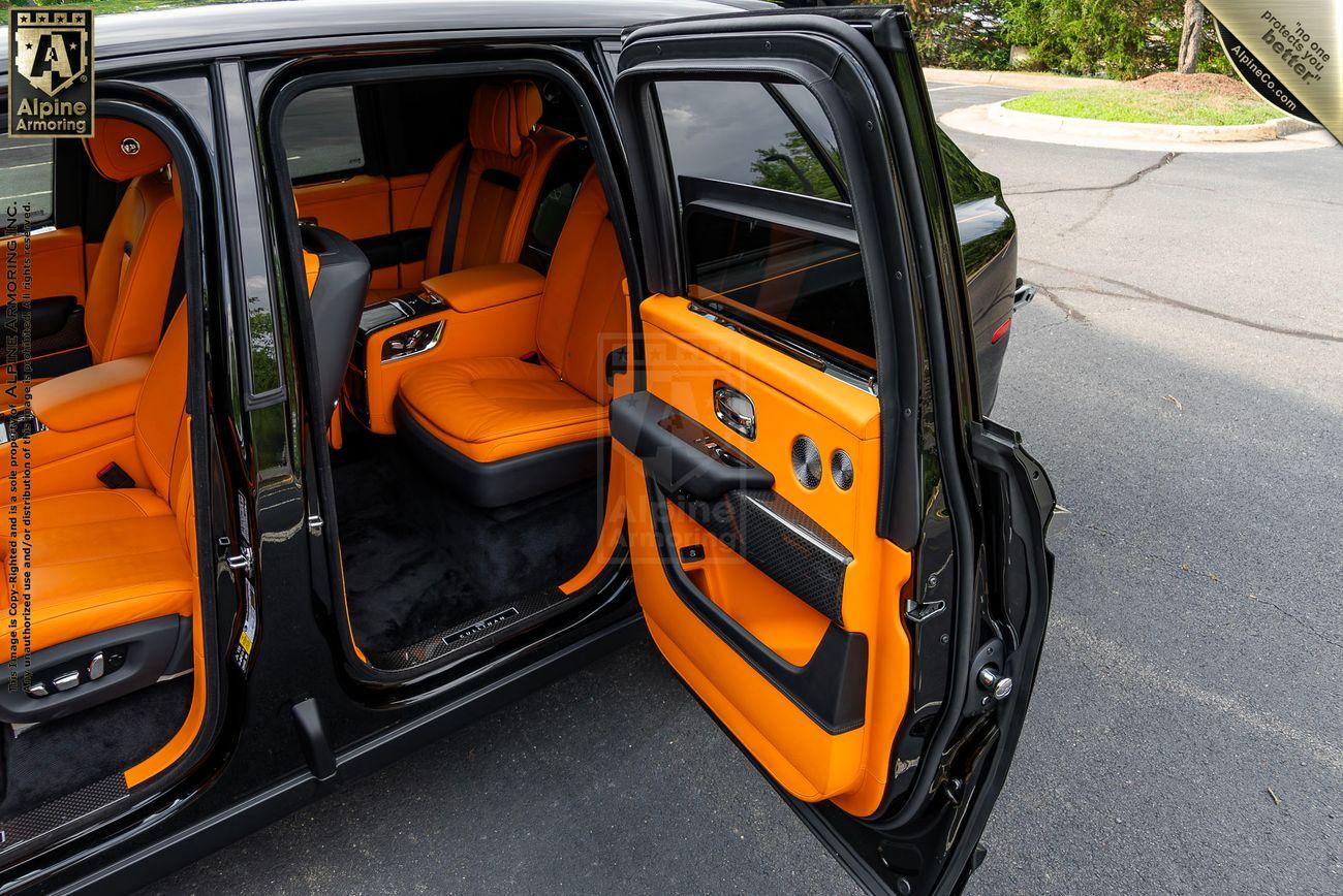 A larmored Rolls-Royce Cullinan SUV with open rear doors reveals an interior featuring vibrant orange seats and accents, a central console with controls, and black flooring. Exterior setting includes pavement and greenery.