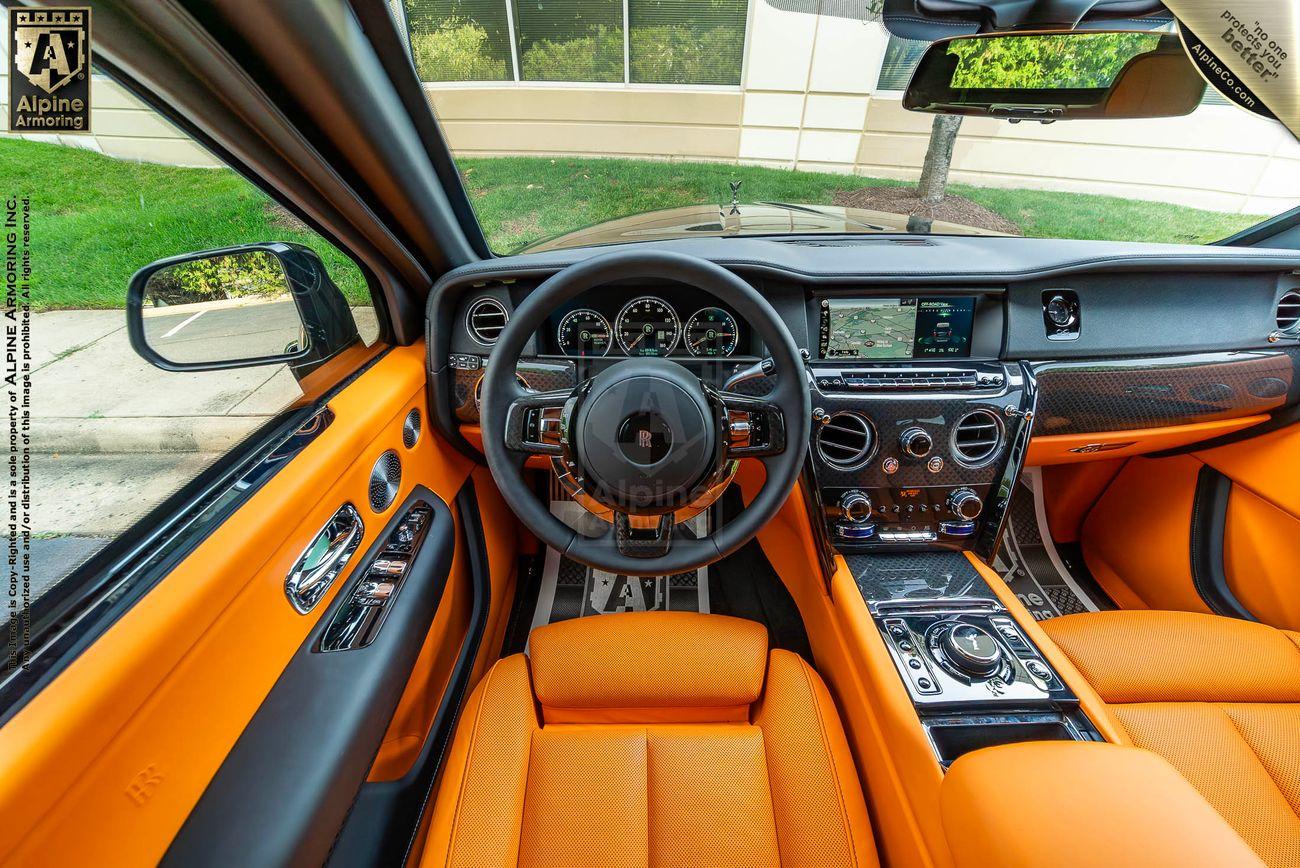 Interior view of an armored Rolls-Royce Cullinan SUV with orange leather seats, a leather-wrapped steering wheel, a touchscreen infotainment system, and a polished central console. Outside, a green lawn and a building are visible.