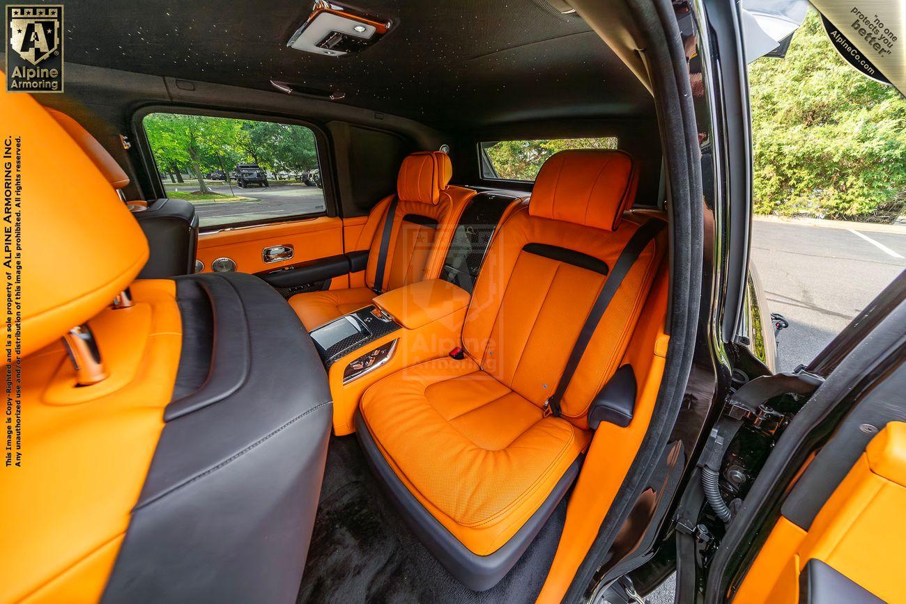 The interior of an armored Rolls-Royce Cullinan with black and bright orange leather seats, a central control console, sunlight streaming through the windows, and trees visible outside.