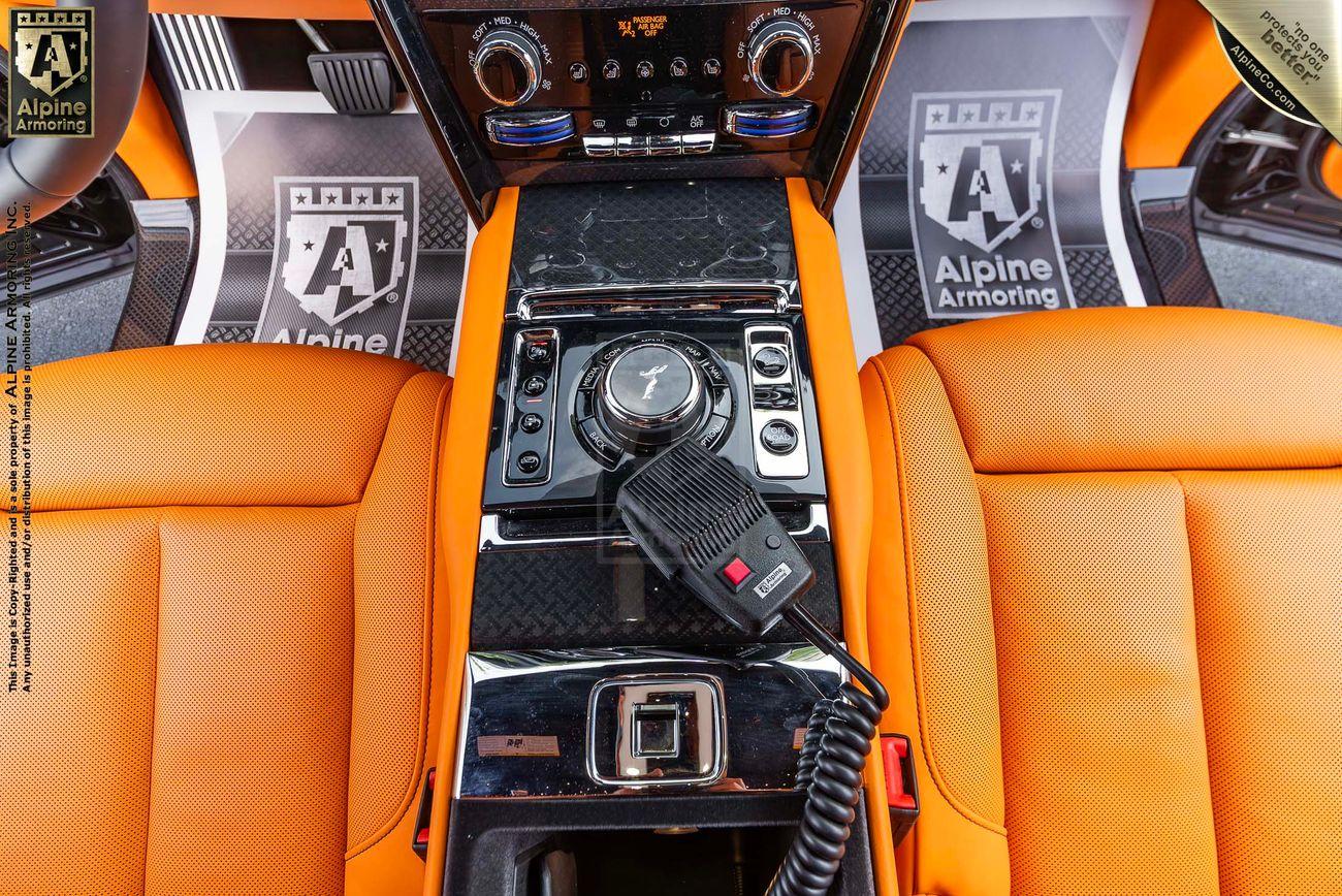 An armored Rolls-Royce Cullinan interior featuring vibrant orange leather seats, a central console with various controls, an Alpine Armoring badge, and a mounted communication device.