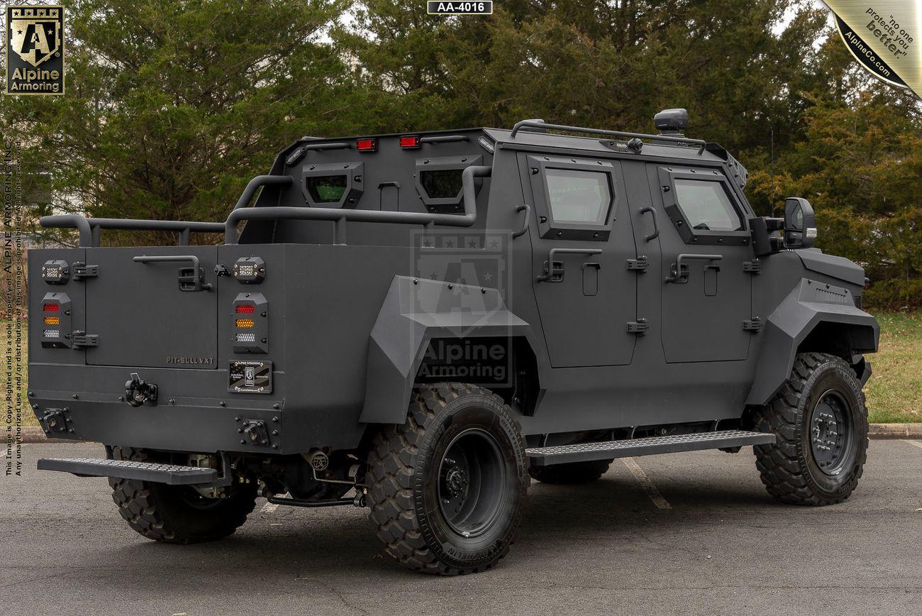 A black armored Pit-Bull VXT is parked on a road with trees in the background. The vehicle features rugged tires, heavy-duty armor, tinted windows, and various modifications for security purposes.