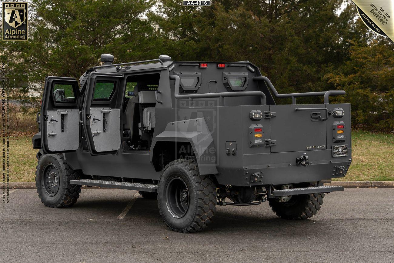 A black, armored Pit-Bull VXT with open doors, parked on a paved surface with trees in the background.