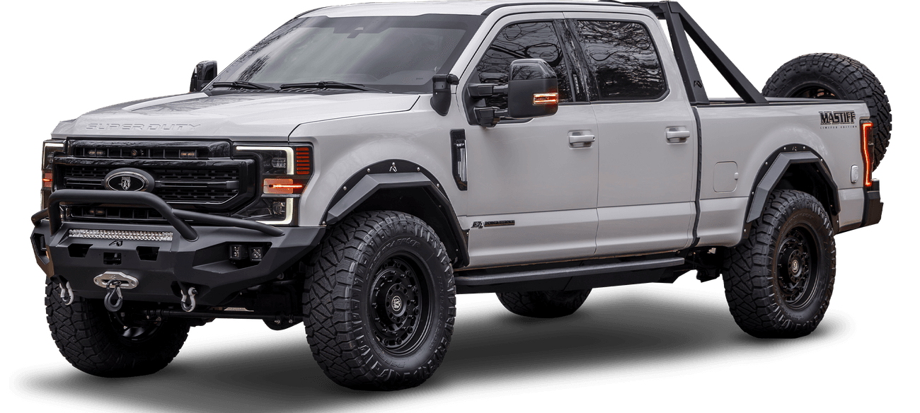 White, four-door armored Mastiff truck with black trim, bull bar, roof rack, and off-road tires. Truck has a mounted spare tire on the rear and side steps.