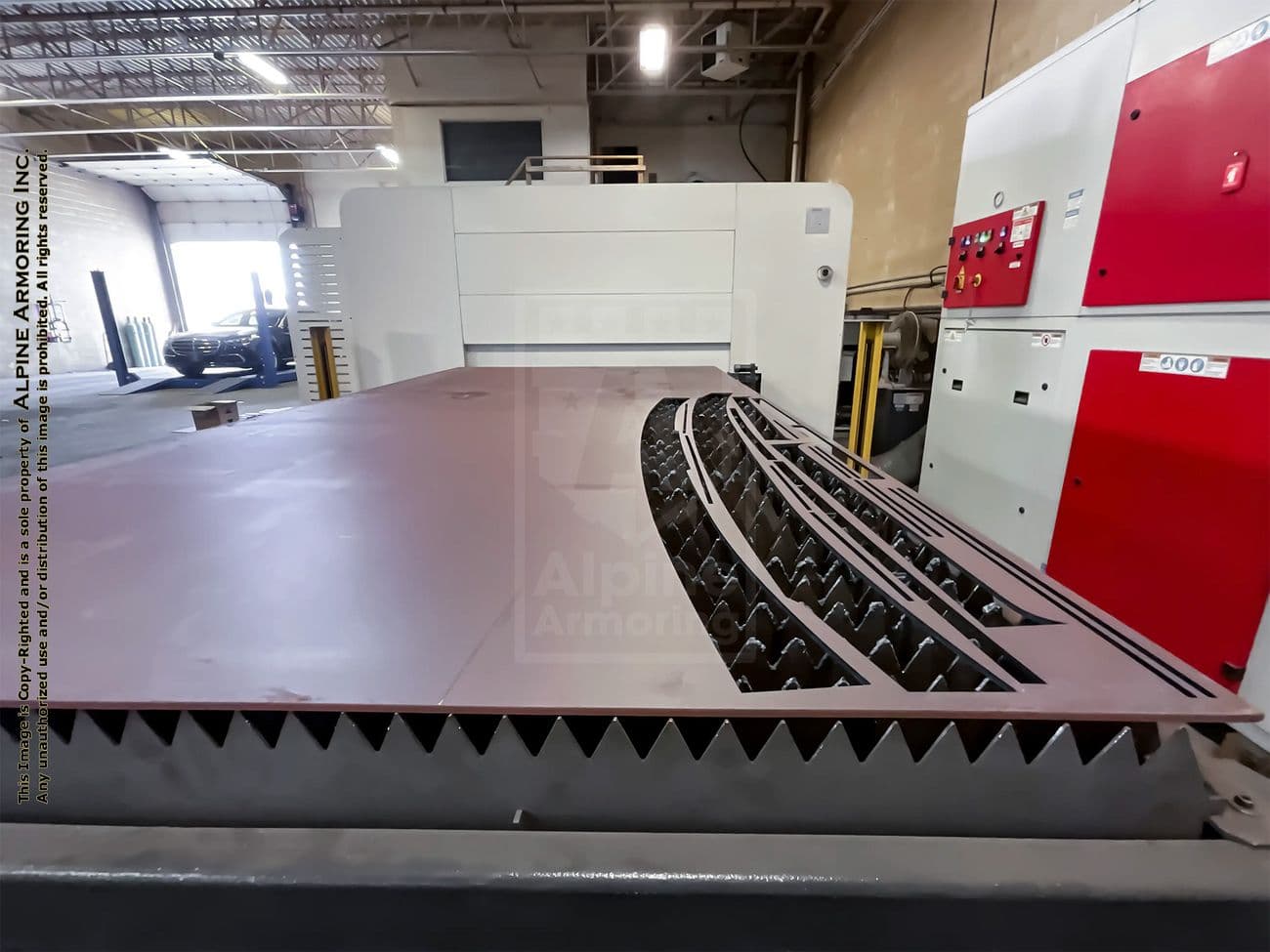 Large industrial laser cutting machine in a workshop setting. Metal sheet on the machine bed with intricate cut patterns. Background includes additional equipment and workshop area.