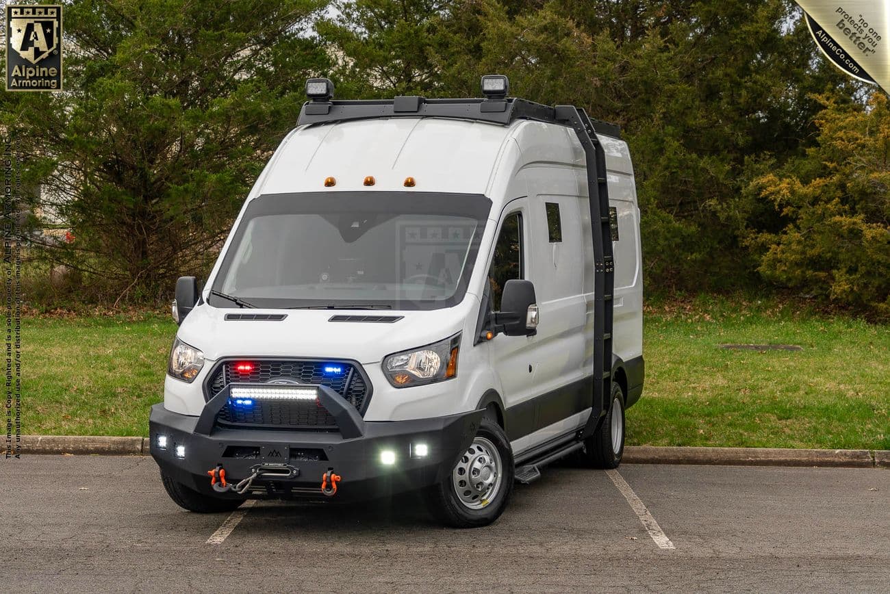 A white armored Pointer van, from Alpine Armoring, equipped with rooftop equipment, a winch, and police lights, parked in a lot with trees in the background.