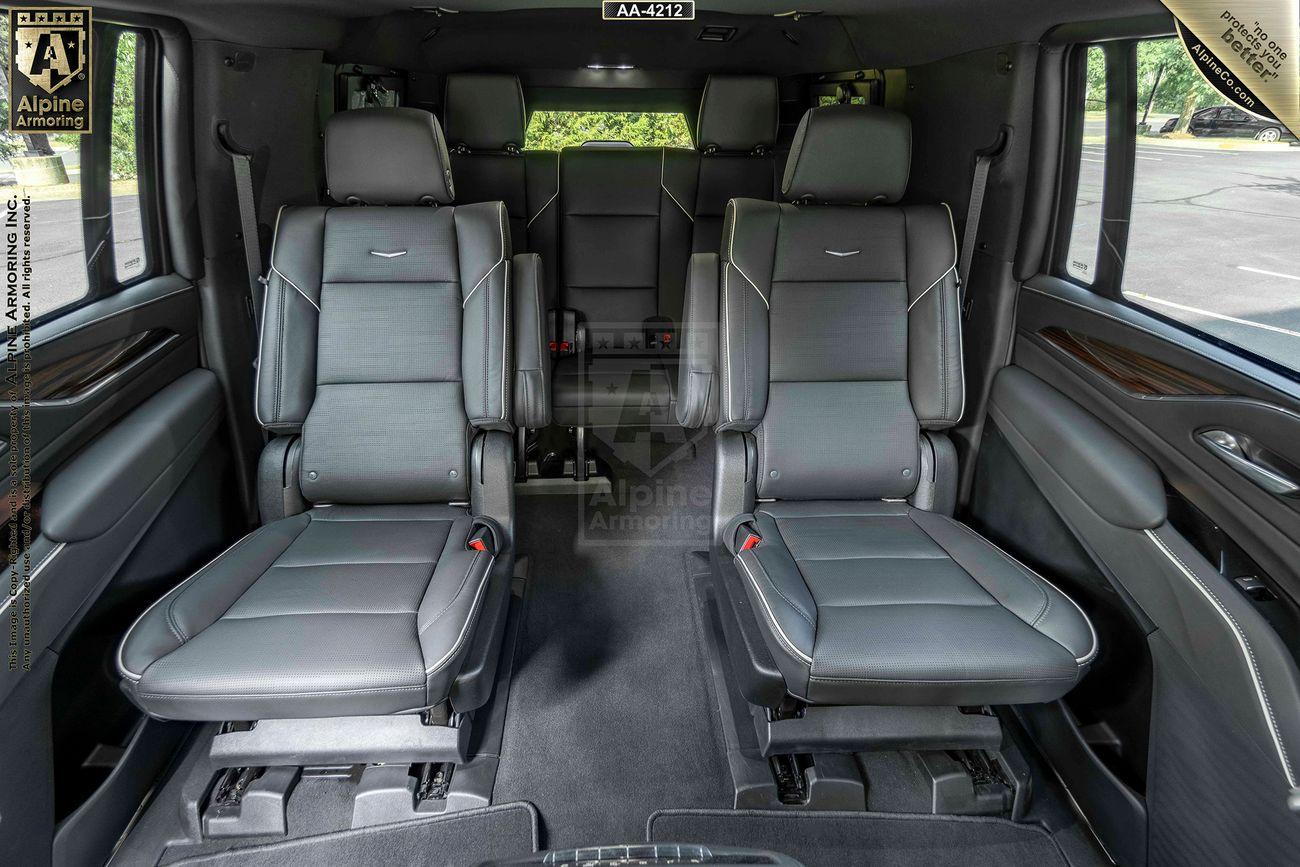 Interior view of a bulletproof Cadillac Escalade ESV SUV showing four leather seats, with two in front and two in the rear, separated by an aisle. The seats are black with detailed stitching, and the windows reveal a green outdoor scene.