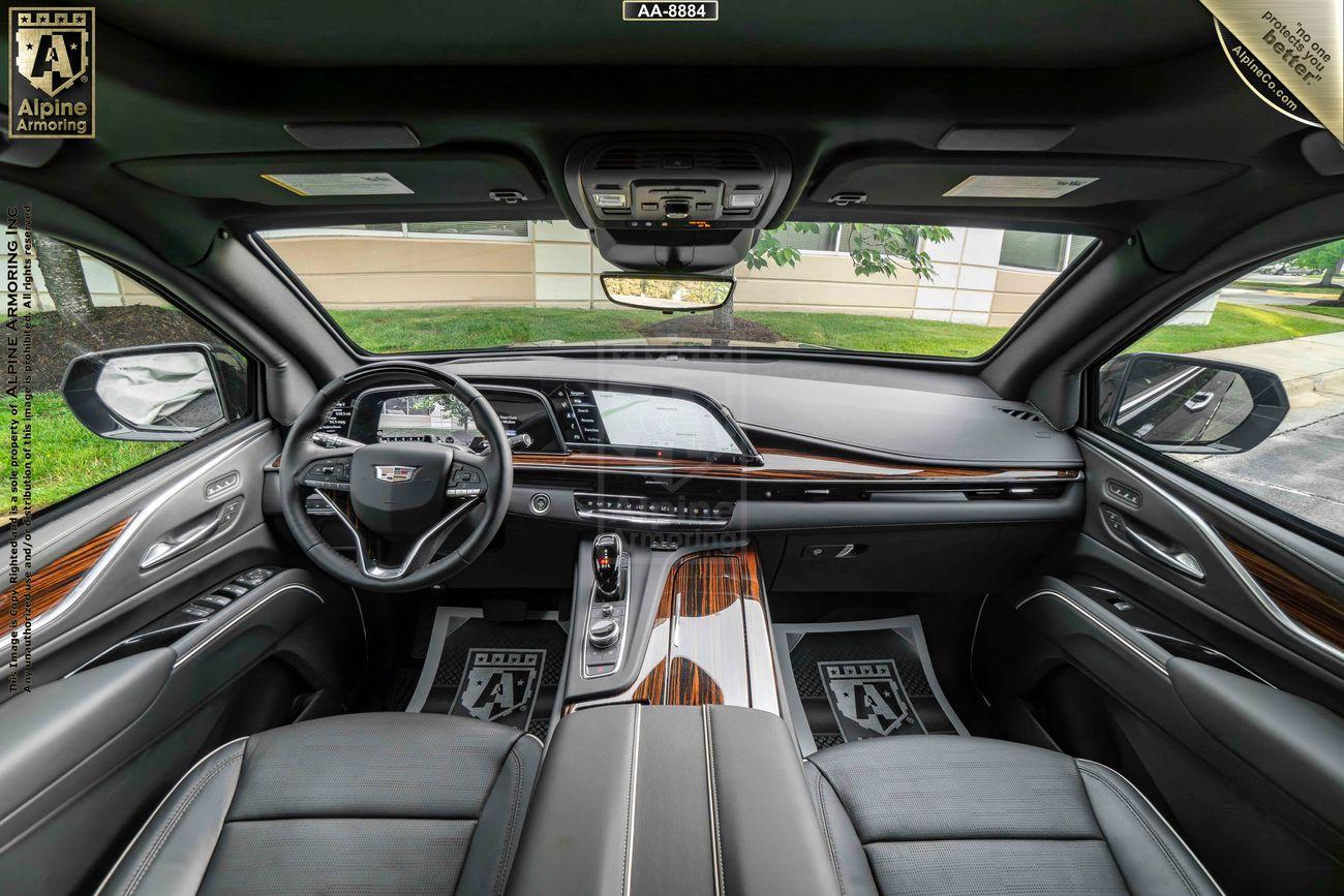 Interior of a  Cadillac Escalade Premium Luxury SUV showing the steering wheel, dashboard with digital displays, wood paneling, leather seats, and various control buttons.