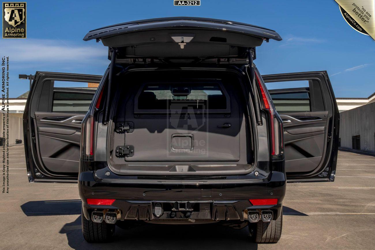 A black armored Cadillac Escalade ESV SUV is shown from the rear with its trunk and back doors open, displaying an empty cargo space. The vehicle is parked in a paved outdoor area under a clear blue sky.