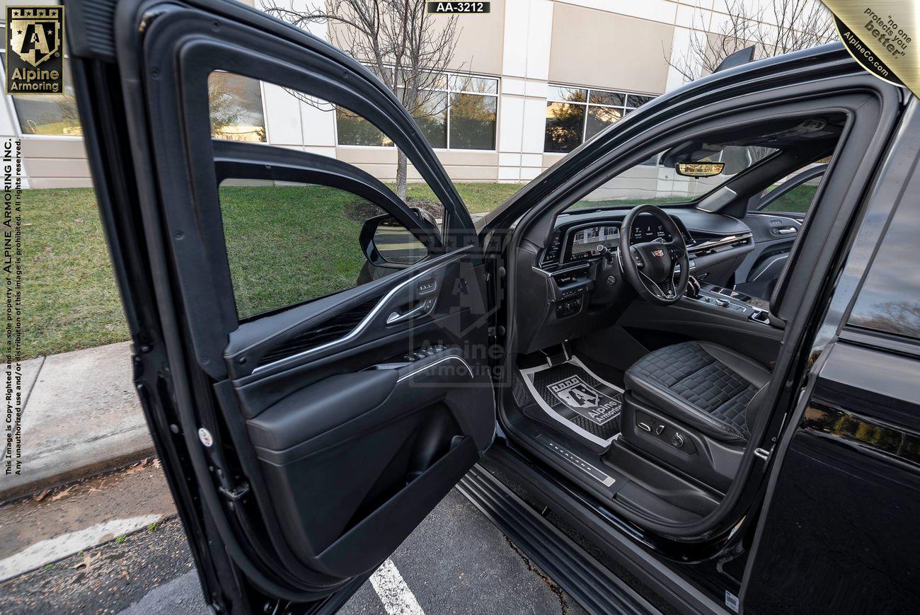 Open door view of a black armored Cadillac Escalade ESV SUV showcasing the front interior, including the dashboard, driver's seat, and custom mats, with a building exterior and grass in the background.