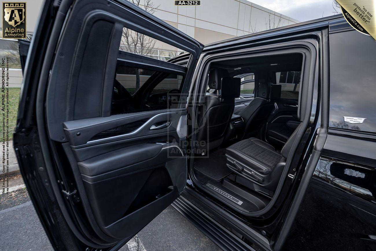 An open rear passenger door of a black bulletproof Cadillac Escalade ESV SUV reveals a spacious interior with two leather seats. Part of the exterior of a building is visible in the background.