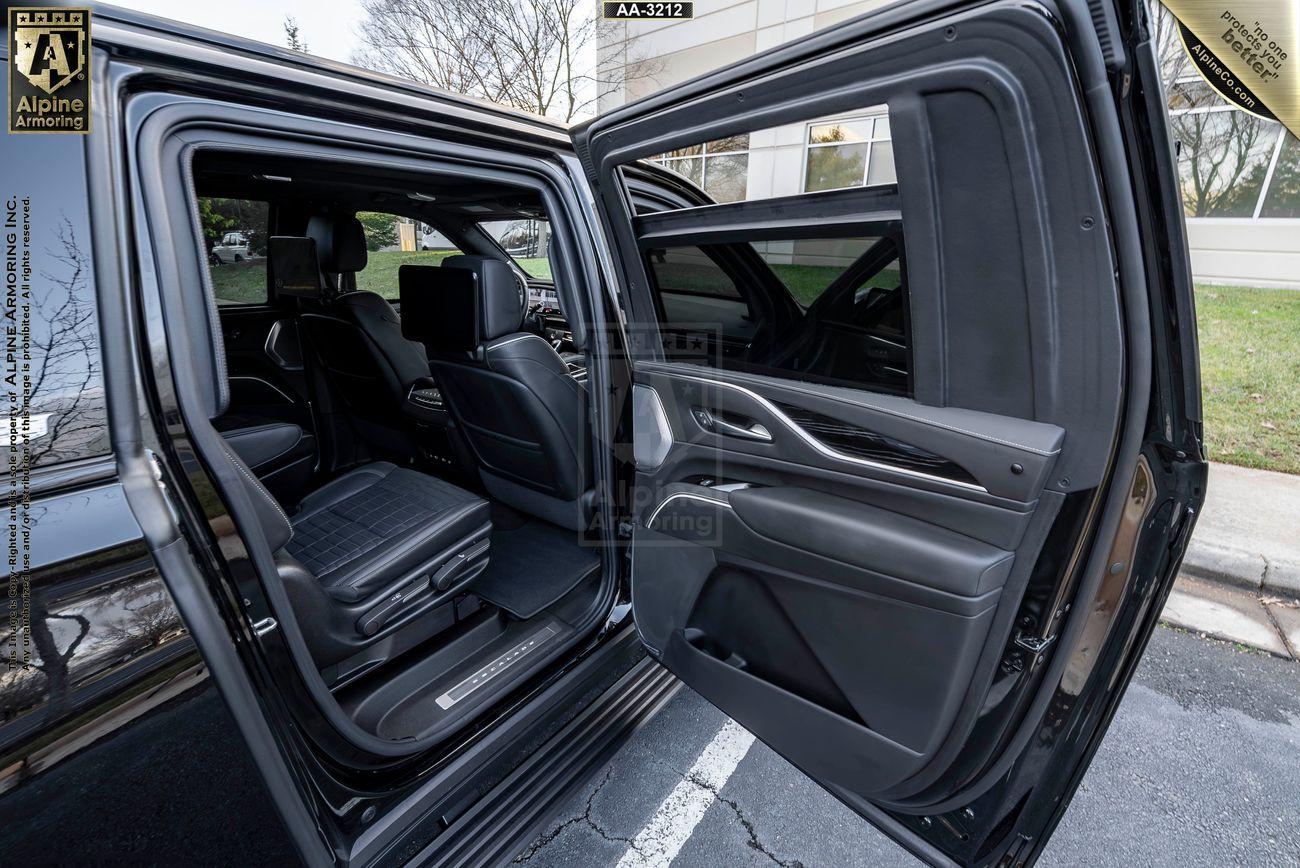 An open backseat of a black bulletproof Cadillac Escalade ESV SUV with two passenger seats, a display screen on the back of each front seat, and a view of the exterior landscape outside the vehicle.