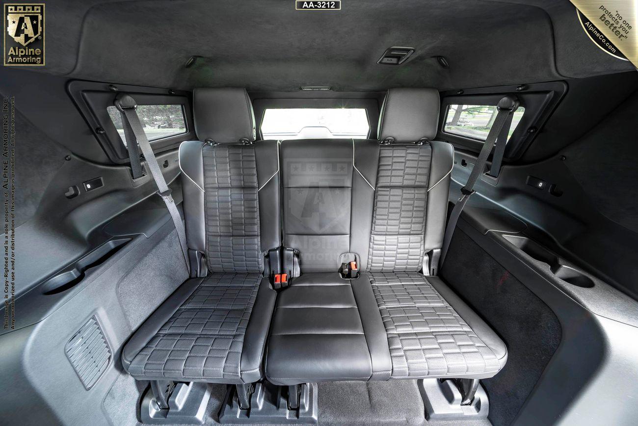 Interior of an armored Cadillac Escalade ESV SUV with three black leather back seats, featuring seat belts and a central armrest. The seats are surrounded by dark paneling and windows.