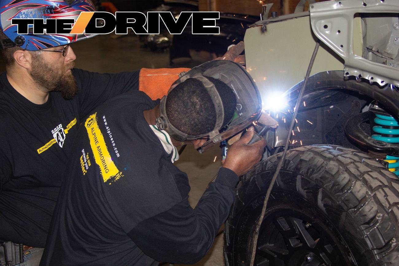 Two Alpine Armoring welders in protective gear work on a vehicle in a garage. One welder, wearing a helmet and gloves, uses a welding torch while the other assists. Sparks fly from the welding point. A logo that reads "THE DRIVE" is visible .