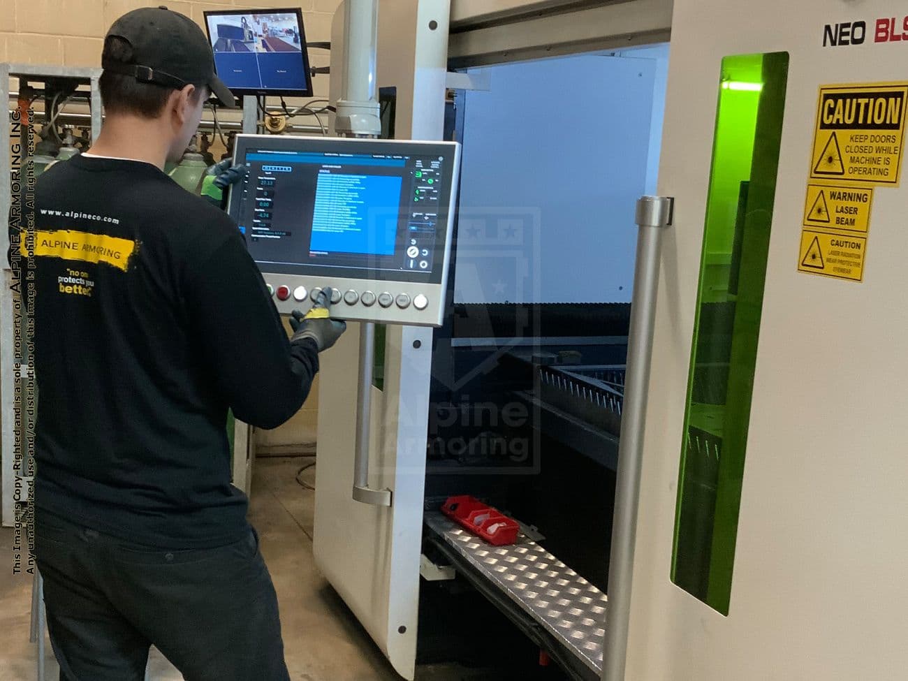 An Alpine Armoring employee operates an industrial machine with a touchscreen interface. They are standing in a workshop with various equipment and two monitors overhead. Safety signs are visible on the machine.