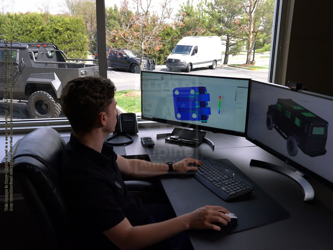 A person is working on a computer with dual monitors displaying 3D design software in a brightly lit office with windows overlooking a parking lot.