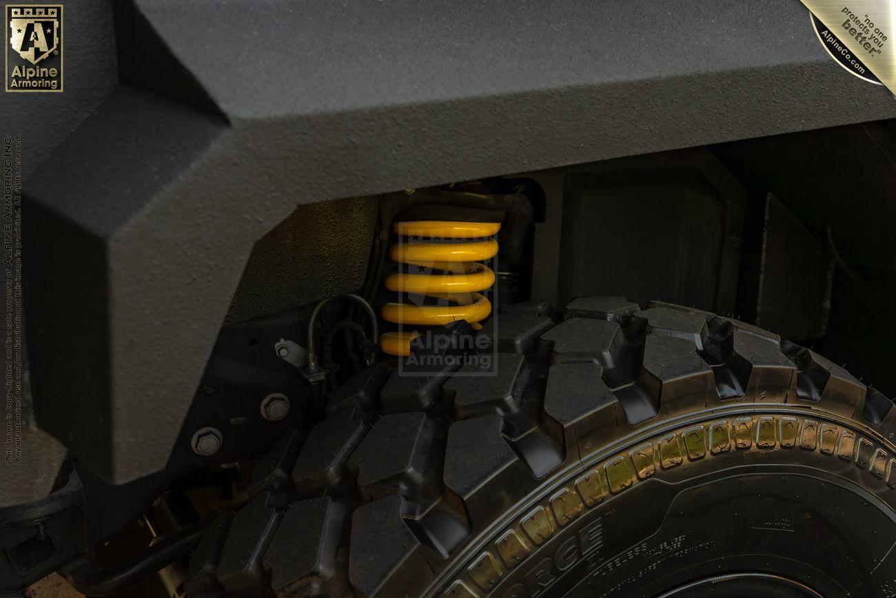 A close-up view of an armored vehicle's wheel well, highlighting the yellow suspension coil and rugged tire from Alpine Armoring.